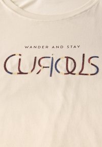 Krämfärgad bomullen t-shirt med rund hals. Text tryckt med flerfärgade bokstäver läser "VANDRA OCH VAR NYFIKEN." Mjuk textur.