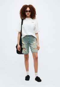 Νεαρή γυναίκα με σγουρά μαλλιά που φοράει μαύρα γυαλιά ηλίου, άσπρο φαρδύ μπλουζάκι, σκισμένο τζιν σορτς, άσπρες κάλτσες, μαύρα παπούτσια χωρίς κορδόνια και μαύρη τσάντα ώμου.