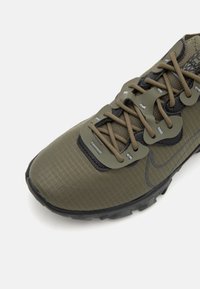 Olivově zelená teniska Nike s texturovaným svrškem, plochými tkaničkami a kontrastními černými detaily. Má polstrovanou podešev a výrazný vzorek na podrážce.