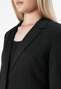 Detailní záběr na ženu v upraveném černém saku přes černý žebrovaný top, ukazující límec a horní část hrudníku.
