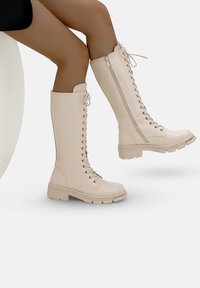 Stivali beige alti fino al ginocchio realizzati in pelle liscia, con un design di allacciatura frontale, zip laterale e suola in gomma texturizzata.
