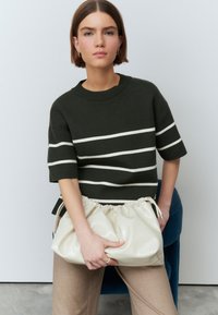 Pulover verde cu dungi, cu guler rotund, asortat cu o geantă crem, cu pliuri. Modelul poartă pantaloni bej, evidențiind un design curat.