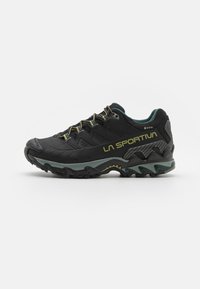 Melnas sporta kurpes ar sietiņu augšdaļu, teksturētu gumijas zoli un zaļiem akcentiem. Iezīme pastiprināta purngalu un GORE-TEX zīmols uz papēža.