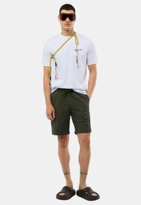 T-shirt bianco con grafica rossa, corda gialla con moschettoni, pantaloni corti verde oliva, calzini beige con testo e sandali neri senza lacci.