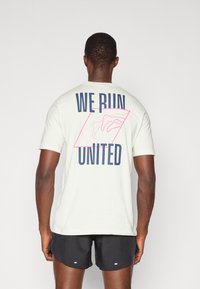 Tricou de culoare deschisă cu guler rotund. Prezintă un text îndrăzneț "WE RUN UNITED" și un design geometric cu contur roz pe spate.