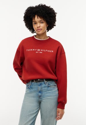 Ung kvinde med krøllet hår iført en rød Tommy Hilfiger sweatshirt og lyseblå jeans mod en hvid baggrund.