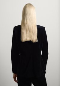 Persona con lunghi capelli biondi lisci che indossa una giacca nera, vista da dietro su uno sfondo chiaro e semplice.