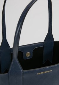 Marineblaue Ledertasche mit strukturierter Oberfläche, ausgestattet mit zwei Griffen und einem magnetischen Druckknopfverschluss. Innenfutter aus schwarzem Wildleder. Sichtbares goldenes Logo.