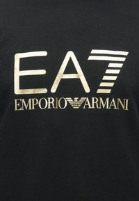 Čierne bavlnené tričko s potlačou zlatého loga: "EA7 EMPORIO ARMANI" so štýlizovanými pásikmi a detailom orlieho emblému.