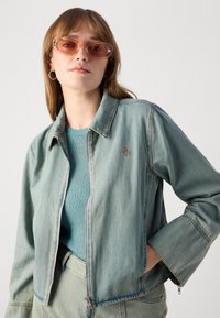 Lyseblå denimjakke med glidelås foran, spiss krage og sidelommer. Båret over en ribbet turkise genser med fremhevede sømmer.
