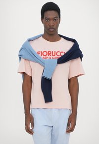 Ανοιχτό ροζ μπλουζάκι φ�τιαγμένο από βαμβάκι με την επιγραφή "Fiorucci Cash & Carry" σε κόκκινο χρώμα, συνδυασμένο με μπλε και ναυτικές πλεκτές ζακέτες, και ριγέ παντελόνια.