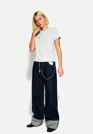 Graues Baumwoll-T-Shirt kombiniert mit weit geschnittenen dunkelblauen Jeans, die einen umgeschlagenen Saum in Hellgrau aufweisen. Kopfhörer hängen von dem Shirt.