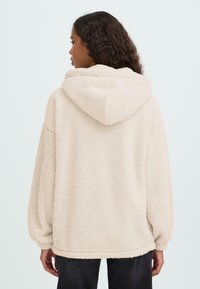 Béžová bunda s kapucí z umělé kožešiny s měkkou strukturou, oversized střih a kapuce. Elastické manžety a lem, minimální detaily švů.