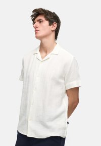 Camisa de manga corta, con botones, en tejido texturizado color crema, con cuello con muesca y sutiles rayas horizontales. Presenta botones en la parte frontal y aberturas laterales.