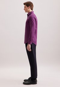 Uomo in profilo che indossa una camicia a maniche lunghe viola con bottoni, pantaloni neri e scarpe da uomo nere, su uno sfondo neutro.