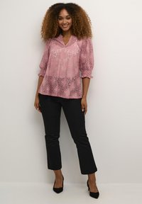 Blouse en dentelle rose avec des motifs floraux, décolleté en V et manches trois-quarts. Associée avec un pantalon noir ajusté et des escarpins noirs.