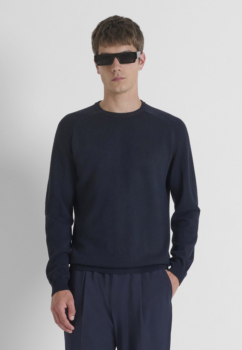 Marineblauwe trui met geribbelde schouderaccenten en ronde halslijn, gemaakt van een zachte brei stof. Gepaard met zwarte rechthoekige zonnebril.