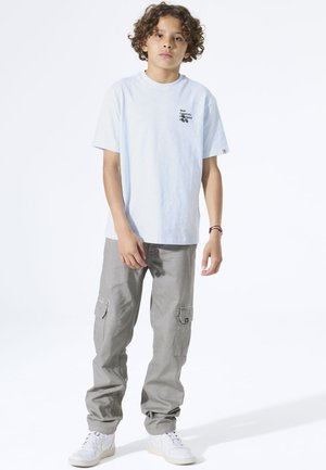 Hellblaues Baumwoll-T-Shirt mit kurzen Ärmeln, versehen mit schwarzem Text und grafischem Design. Kombiniert mit grauer Cargohose und weißen Turnschuhen.