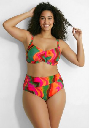 Uśmiechnięta kobieta z kręconymi włosami pozuje w kolorowym bikini z wysokim stanem z pomarańczowymi, różowymi i zielonymi zawijasami na białym tle.