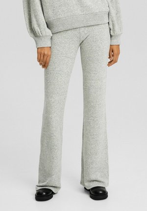 Person trägt hellgraue, gestrickte Hosen mit weitem Bein und passenden Pullover, steht mit locker an den Seiten hängenden Händen, schwarze Schuhe sichtbar.