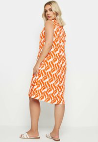 Oranssi ja valkoinen hihaton mekko, jossa on aalto kuvio, polvimittainen, valmistettu kevyestä kankaasta. Malli, joka käyttää valkoisia sandaaleja, seisoo sivuttain.