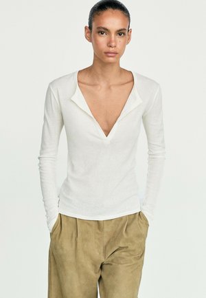 Donna con capelli scuri raccolti che indossa una maglia bianca a maniche lunghe con scollo a V profondo e pantaloni larghi beige con le mani in tasca.