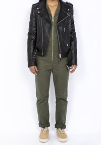 Svart läderbikerjacka med silverdetaljer, olivgrön jumpsuit under, och beiga mockasneakers. Avslappnad och funktionell design.