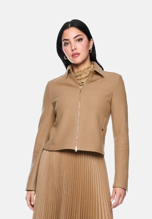 Woman with dark hair wearing gold hoop earrings, a beige zip-up jacket, a patterned beige turtleneck, and a pleated beige skirt.