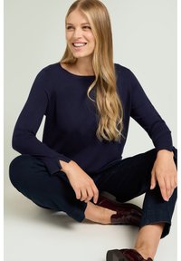 Maglione lavorato a maglia di colore blu navy con maniche lunghe e scollo arrotondato, abbinato a pantaloni scuri e scarpe lucide color bordeaux, seduto su una superficie chiara.