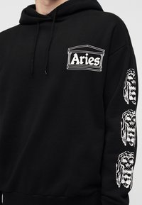 Μαύρο φούτερ με λευκό λογότυπο "Aries" μέσα σε περίγραμμα κλασικού κτιρίου στο στήθος και τρία λευκά σχέδια κρανίων στο δεξί μανίκι.