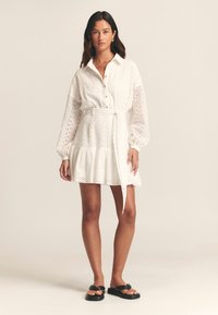 Bílé šaty košilového stylu se zdobením z průsvitného materiálu, dlouhými nabíranými rukávy, zapínáním na knoflíky vpředu a opaskem v pase. Volná, vrstvená sukně. Černé sandály.