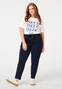 Witte T-shirt met blauwe "AMOUR" opdruk, gecombineerd met navy tapering broek met een drawstring taille en zijzakken; compleet met beige sneakers.