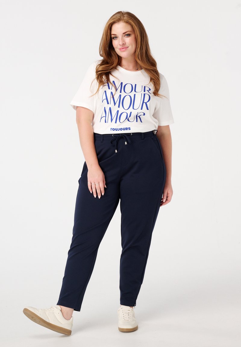 Witte T-shirt met blauwe "AMOUR" opdruk, gecombineerd met navy tapering broek met een drawstring taille en zijzakken; compleet met beige sneakers.