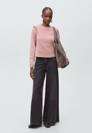 Junge Frau in einem rosa Langarmoberteil, weiten schwarzen Jeans, die eine große graue Wildleder-Schultertasche trägt, vor einem neutralen Hintergrund stehend.