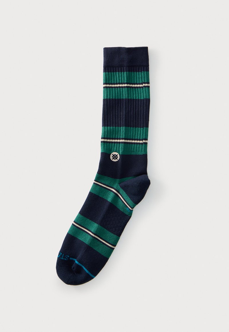 Chaussette bleu foncé avec des rayures horizontales vertes et blanches et un petit logo circulaire près de la cheville, posée à plat sur un fond clair.