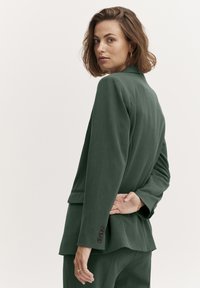 Tumši zaļš piegriezuma bleizeris ar ieliektām apmalēm, trīs pogām uz piedurknēm un gludu tekstūru. Modelis pozē, ar roku uz muguras.