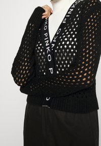 Svart stickad kofta med diamantformade utskärningar, utrustad med en märkespräglad V-ringning och ribbad nederkant. Designad för att bäras över olika toppar.