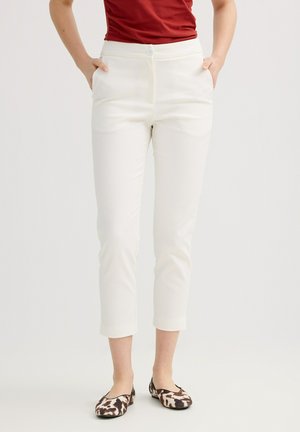 Persona con pantalones blancos recortados, camiseta roja y zapatos planos marrones y blancos con estampado, de pie con las manos en los bolsillos.