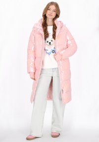 Młoda kobieta ubrana w błyszczącą jasnoróżową długą puchową kurtkę, białą bluzę z grafiką psa, jasnoniebieskie jeansy o szerokich nogawkach oraz różowe trampki