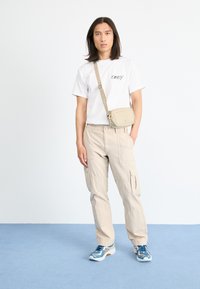 Beige cargo nohavice, biele tričko s logom "OBEY" a modré športové topánky. Malá béžová crossbody taška. Jednoduchý dizajn, uvoľnený strih.