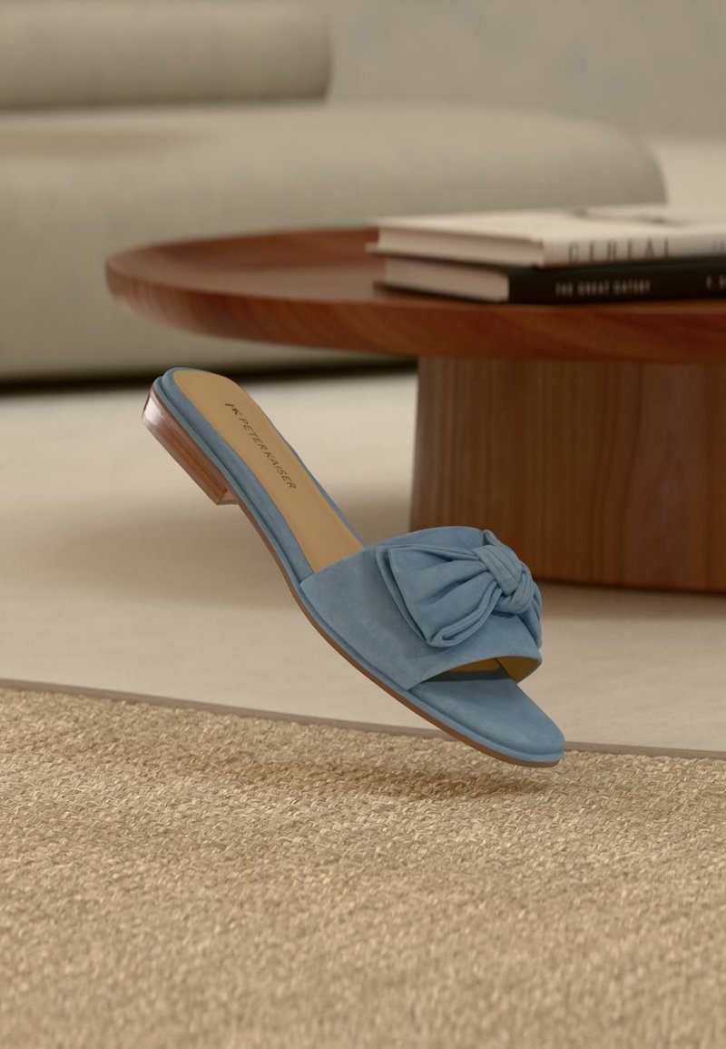 Sandale mule en daim bleu à bout ouvert avec un détail nœud, posée sur un tapis beige texturé près d'une table basse en bois avec des livres empilés.