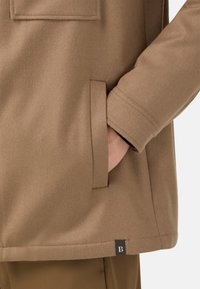 Brun ullblandad kappa med sidoficka, synliga sömmar och en logotagg vid nederkanten. Tyget har en mjuk, texturerad finish.