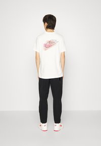 Camiseta blanca de Nike con un logotipo gráfico en rojo y rosa en la parte posterior, combinada con pantalones negros y zapatillas blancas con acentos rojos.