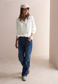 Chemisier blanc à manches longues avec un petit plastron, jeans en denim bleu et baskets claires, complété par une casquette beige, se tenant sur une surface neutre.