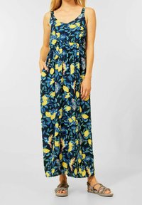 Maxi šaty z tmavě modré látky zdobené žlutými citrony a květinovými vzory; mají tenké ramínka a boční kapsy, doplněné o šedé sandály.