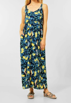 Maxi-jurk van donkerblauwe stof versierd met gele citroenen en bloempatronen; heeft spaghettibandjes en zijzakken, gecombineerd met grijze sandalen.