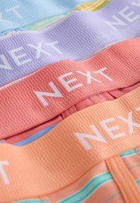 Ceintures élastiques colorées empilées sur des sous-vêtements, chacune avec le nom de la marque "NEXT" en texte blanc, dans des tons pastel violet, corail, pêche et bleu.