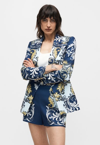 Γυναίκα με σκούρα μαλλιά που φοράει ένα μπλε, λευκό και χρυσό σακάκι με σχέδια και αντίστοιχα σορτς, με σταυρωμένα χέρια, που στέκεται μπροστά σε απλό φόντο.