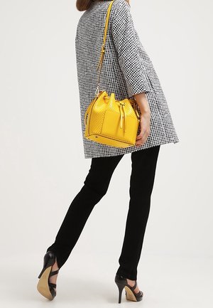 Sac seau en cuir jaune avec texture perforée et fermeture à cordon, associé à une tunique à carreaux noirs et blancs et un pantalon noir.