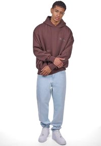 Brun oversized hættetrøje med et broderet logo, kombineret med lyseblå jeans og hvide sneakers, der har en afslappet pasform og manchetter.